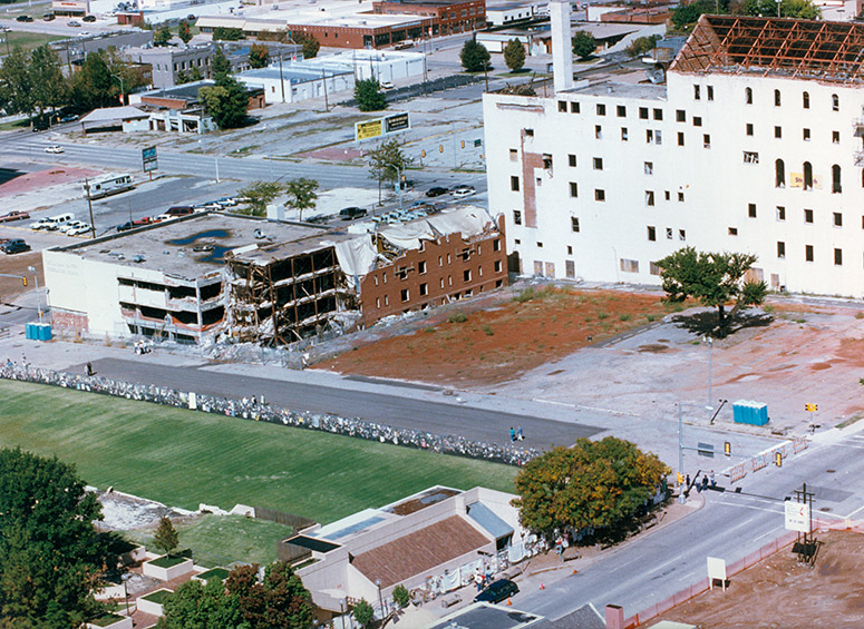 The grass to the left was the former footprint of the Alfred P. Murrah Federal Building. Above that, the original fence is visible, along with Fifth street – now the Reflecting Pool – the Oklahoma Water Resources Board and Athenian buildings, and the Survivor Tree to the right.
